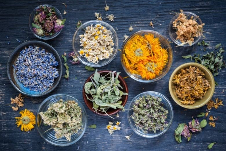 Assorted dried herbs and flowers in bowls on a dark wooden surface, used in acupuncture treatments.