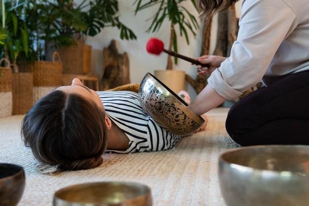 A woman lies on a carpet with her head resting in a large singing bowl while another person holds a mallet nearby, involved in a sound therapy session.