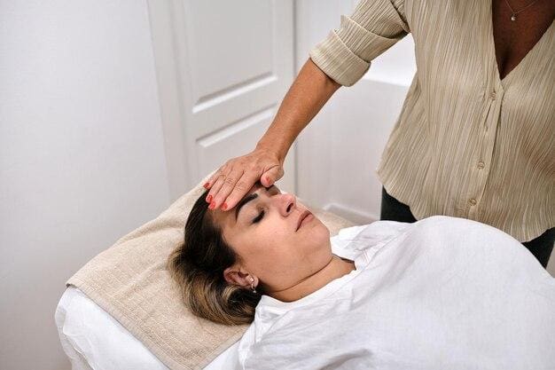 A woman lying down receiving a forehead massage from another woman in a serene, clinical setting.