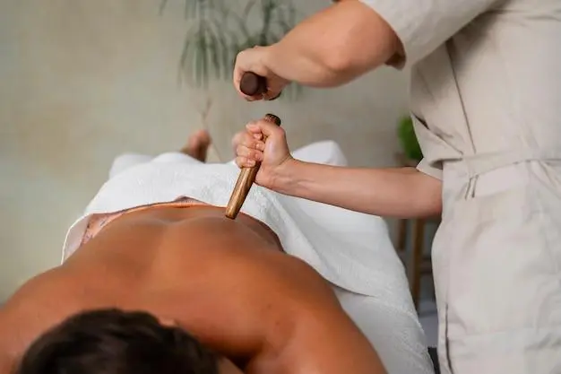 Person receiving a back massage with a wooden tool by a professional masseuse, lying face down on a massage table covered with a white towel.