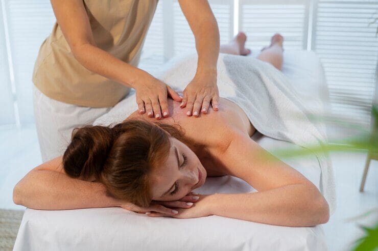 A person is lying face down on a massage table with a towel covering the lower body while receiving a back massage from a masseuse with hands on the person's back.