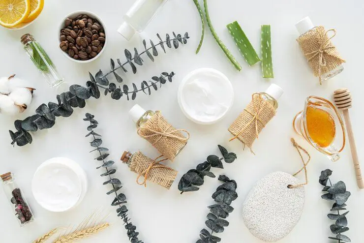 A flat lay of skincare items including jars of cream, bottles with burlap, eucalyptus, aloe vera, honey, coffee beans, essential oils, cotton, and a pumice stone on a white background.