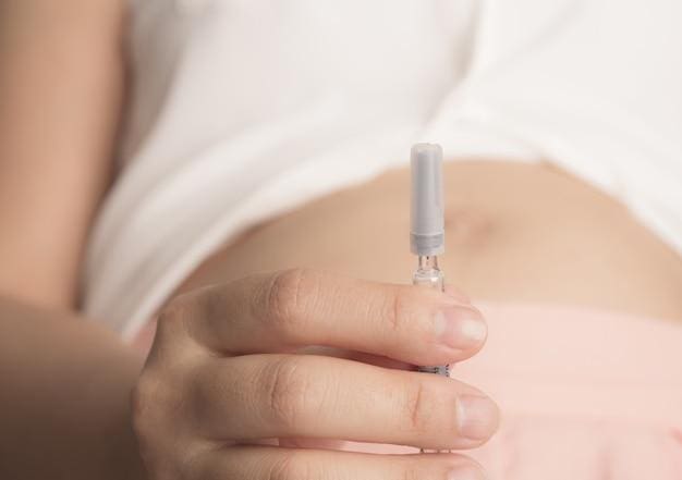 Close-up of a person holding a capped syringe near their bare abdomen. The individual is wearing a white top and pink bottom, and the focus is on the hand and syringe.