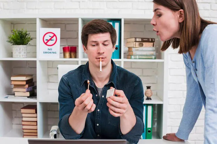 A man is about to light a cigarette in an office with a "No Smoking" sign behind him, while a woman blows air at him disapprovingly.