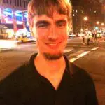 A smiling man with short hair and a goatee stands at an urban street corner at night, with city lights and a few people visible in the background.