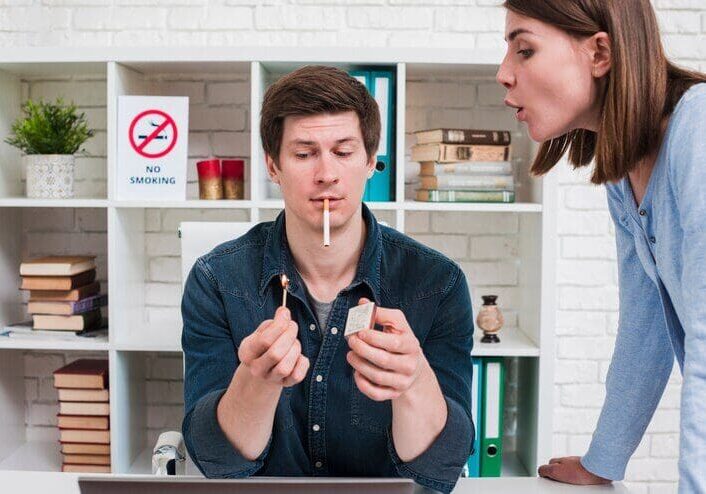 A man is about to light a cigarette in an office with a "No Smoking" sign behind him, while a woman blows air at him disapprovingly.