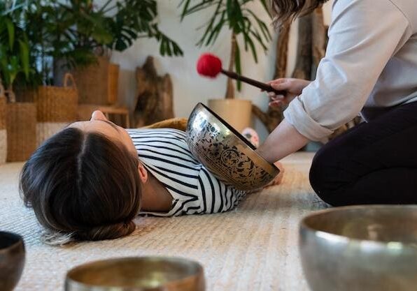 A woman lies on a carpet with her head resting in a large singing bowl while another person holds a mallet nearby, involved in a sound therapy session.