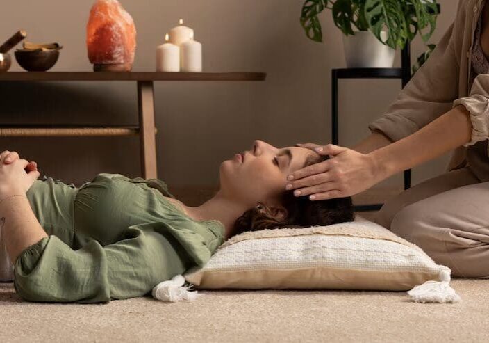 A person lying on a cushion with closed eyes is receiving a head massage from another person in a calm setting with candles and a salt lamp.