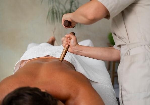 Person receiving a back massage with a wooden tool by a professional masseuse, lying face down on a massage table covered with a white towel.