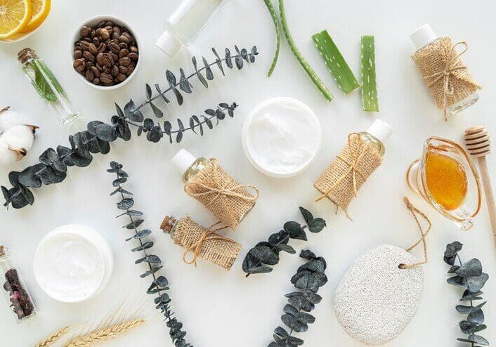 A flat lay of skincare items including jars of cream, bottles with burlap, eucalyptus, aloe vera, honey, coffee beans, essential oils, cotton, and a pumice stone on a white background.